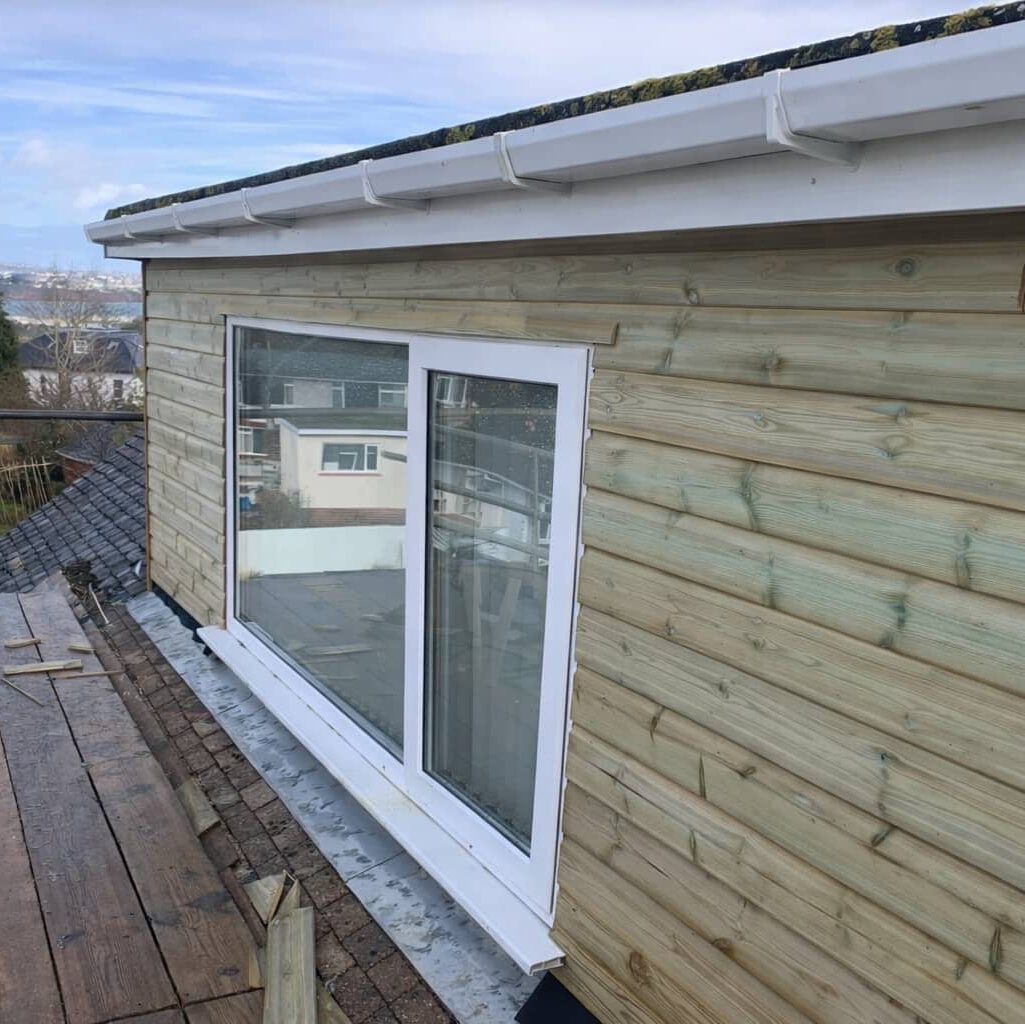 A newly constructed dormer with light wood siding, white trim, and a large sliding window is shown on a sloped roof under a cloudy sky. Scaffolding and nearby houses are visible in the background.