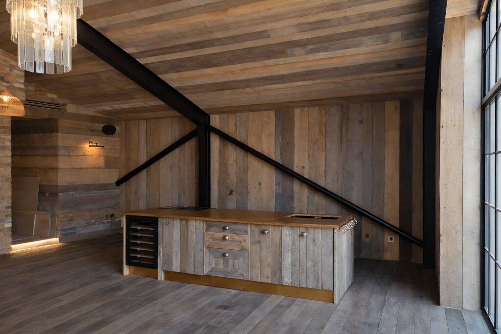 A modern kitchen with rustic wood paneling, an island with a gold countertop, built-in wine rack, and black metal ceiling beams under a sloped ceiling. Large windows let in natural light.