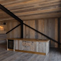 A modern kitchen with rustic wood paneling, an island with a gold countertop, built-in wine rack, and black metal ceiling beams under a sloped ceiling. Large windows let in natural light.