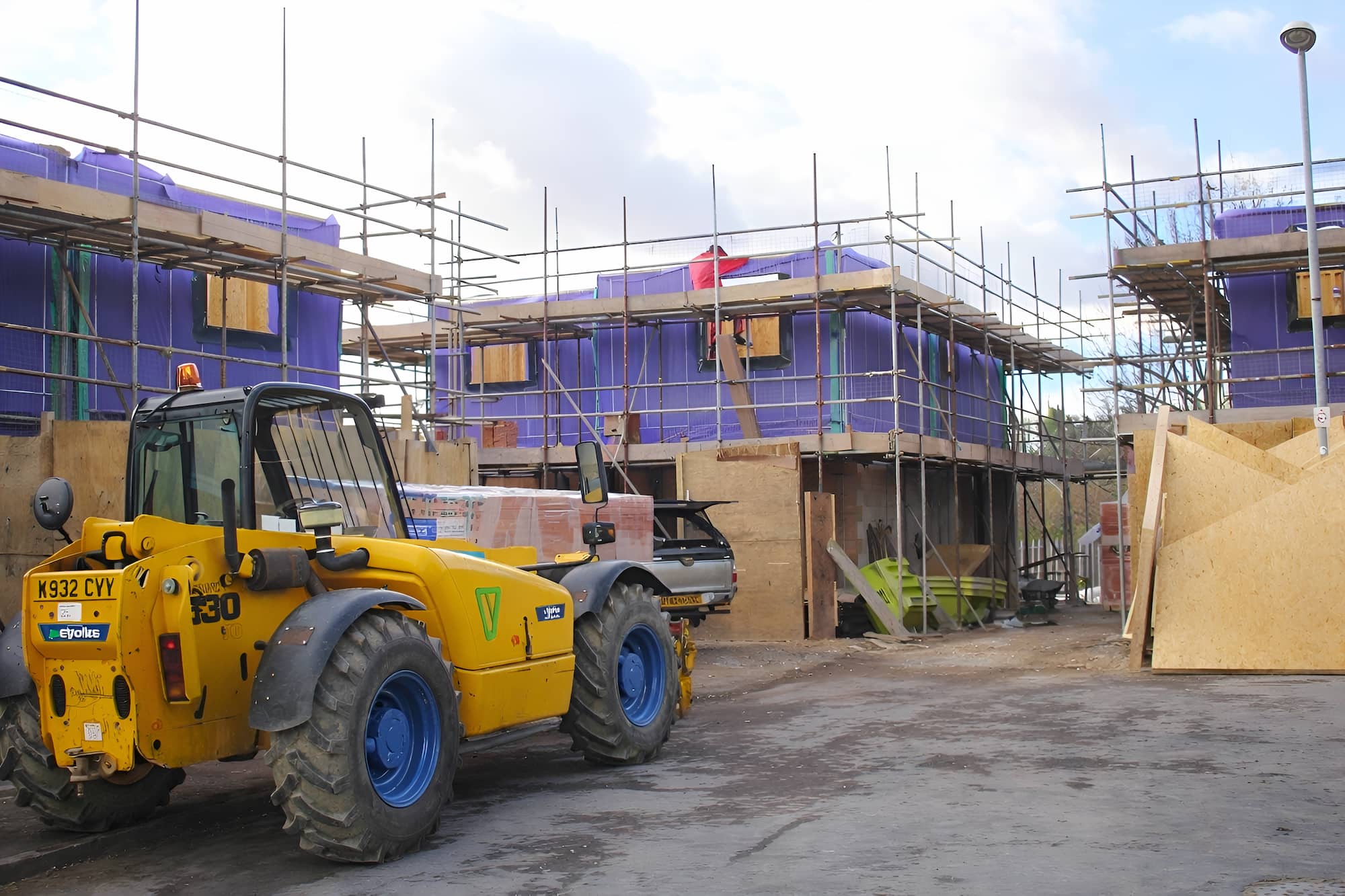 A construction site with partially built houses covered in purple material, surrounded by scaffolding. A yellow forklift is parked in the foreground, and wood panels and building materials are scattered around.
