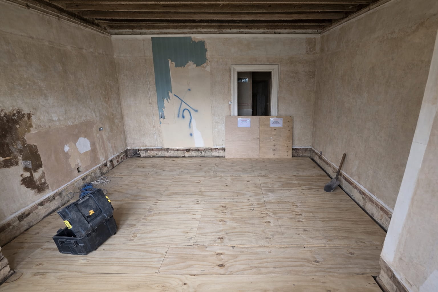 A room under renovation with exposed wooden ceiling beams, bare plaster walls, and plywood flooring. A black toolbox and a broom are on the floor, and two plywood boards partially block the doorway at the far end.