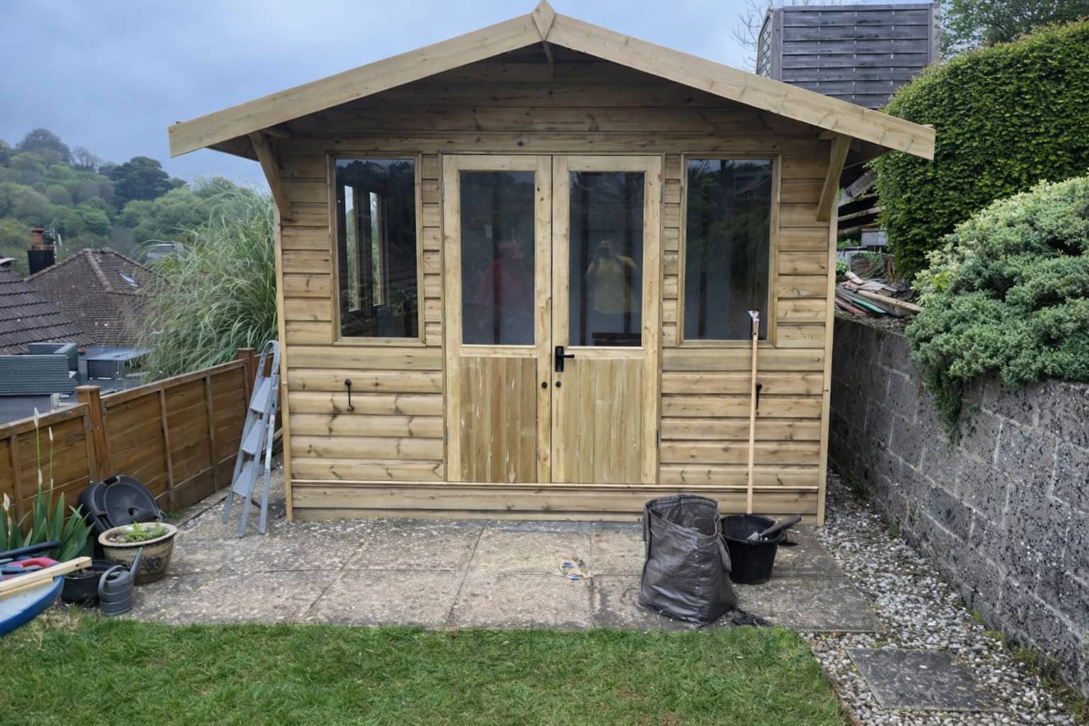 A wooden garden shed with double glass doors stands on a paved patio, surrounded by grass, plants, and gardening tools. The shed is set against a stone wall, with houses and greenery in the background.