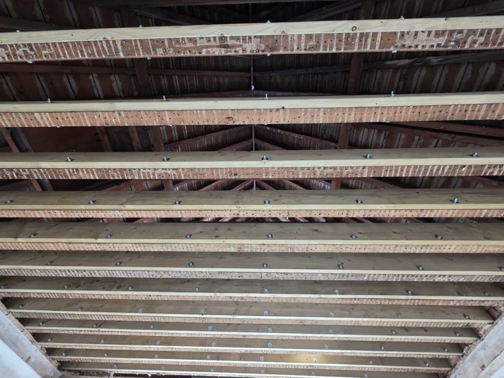 View of an unfinished wooden ceiling with exposed beams, rafters, and metal fasteners. The structure shows no insulation or ceiling panels, revealing the framework of the roof above.